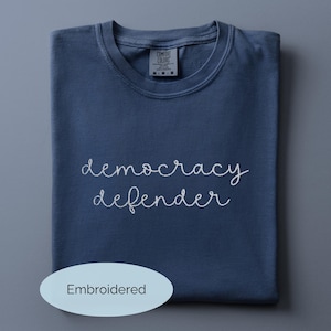 May include: A navy blue crew neck t-shirt featuring the white embroidered text "democracy defender" centered on the chest. The garment includes a Comfort Colors brand label at the inner neckline and a light blue oval tag labeled "Embroidered".