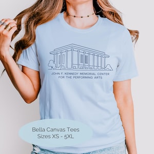 May include: A light blue t-shirt featuring a dark blue line art illustration of the John F. Kennedy Memorial Center for the Performing Arts. The text below the building reads JOHN F. KENNEDY MEMORIAL CENTER FOR THE PERFORMING ARTS. A label on the shirt indicates Bella Canvas Tees Sizes XS - 5XL.