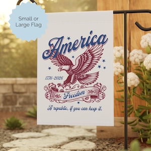 May include: A white garden flag with a patriotic design. The flag features the word "America" in blue script above a red and blue eagle. Below the eagle is a banner with the word "Freedom". The text "1776-2026" and "A republic, if you can keep it" are also included.