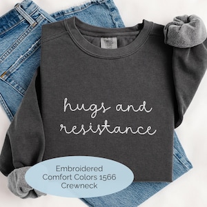 May include: A dark charcoal gray crewneck sweatshirt featuring the white embroidered text "hugs and resistance" on the chest. The garment has a ribbed neckline and cuffs, with a Comfort Colors brand label visible inside the collar area.