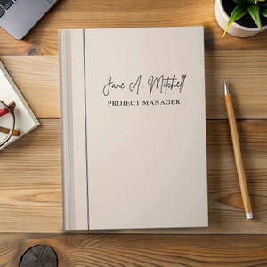 May include: A light beige notebook with a vertical dark line on the left side. The cover features the text Jane A. Mitchell in a script font and the words PROJECT MANAGER in a sans-serif font centered on the front cover of the notebook.