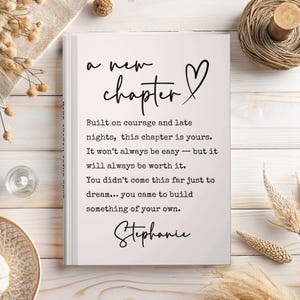 May include: A hardcover book with the words "a new chapter" and a heart symbol in black script. The book contains inspirational text about courage and building something of your own, signed "Stephanie".