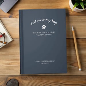 May include: A dark blue journal with the text "Letters to my Dog" in a script font, a paw print, and the phrases "BECAUSE I'M NOT DONE TALKING TO YOU" and "IN LOVING MEMORY OF CHARLIE". A pencil and a plant are visible.