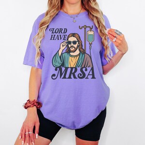 May include: A lilac-colored t-shirt with the text "LORD HAVE MRSA" above an illustration of Jesus wearing sunglasses and an IV drip. The graphic is in shades of green, yellow, and brown.