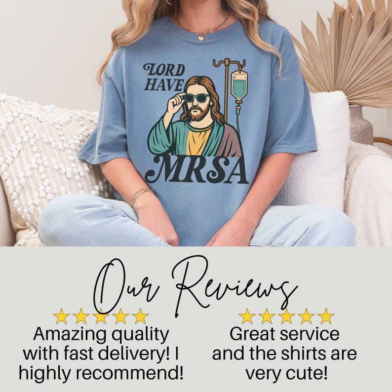 May include: A light blue t-shirt with a graphic of a person with sunglasses and the text "LORD HAVE MRSA." The graphic includes an IV drip bag. The shirt is part of a clothing collection.