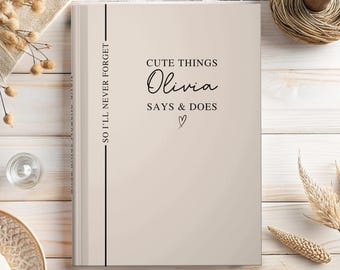 May include: A beige hardcover journal with black text that reads CUTE THINGS Olivia SAYS & DOES with a small heart icon and a vertical line of text on the left side that reads SO I'LL NEVER FORGET. The journal has a clean, neutral design.