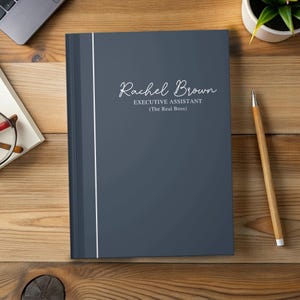 May include: A dark gray notebook with the name "Rachel Brown" in elegant script, along with the title "Executive Assistant (The Real Boss)". A thin white vertical stripe runs along the left side. The notebook is on a wooden surface with a pencil and a plant.