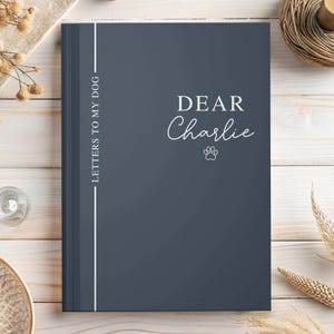 May include: A dark blue journal with the text "DEAR Charlie" in white script and a paw print. The spine has the text "LETTERS TO MY DOG" in white. The journal is on a light wood surface.
