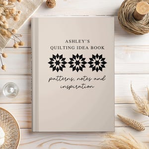 May include: A beige book titled "Ashley's Quilting Idea Book" with black text and geometric star designs. The book cover also includes the words "patterns, notes and inspiration" in a script font. The book is on a light wood surface.