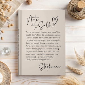 May include: A beige journal with the words "Note to Self" and a heart symbol in black script. The journal contains inspirational text and is signed "Stephanie". The journal is on a light wooden surface.