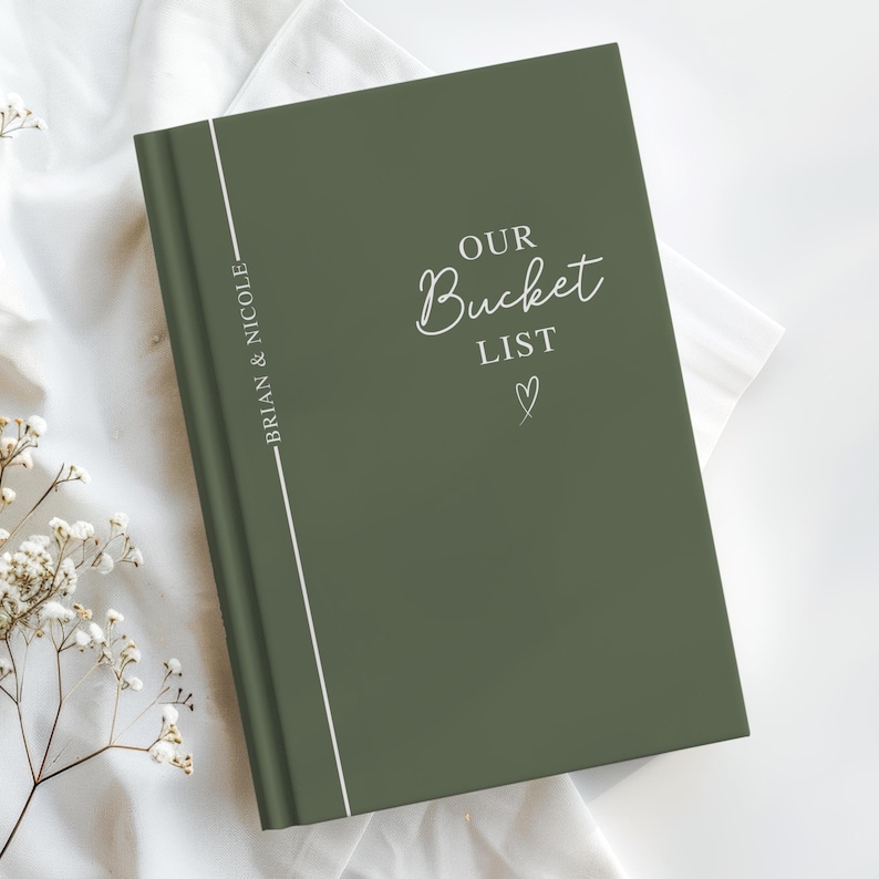 Pu&ograve; includere: Un diario verde con copertina rigida con le parole "OUR Bucket LIST" in corsivo bianco e un piccolo cuore. Il dorso ha il testo "BRIAN & NICOLE" in verticale. Una linea verticale bianca corre lungo il dorso. Il diario &egrave; su una superficie bianca con piccoli fiori bianchi.