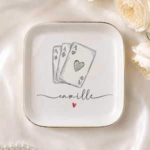 May include: A square white ceramic trinket dish with a gold-toned rim. The center features a black line-art illustration of three playing cards showing aces, with the name "camille" written in black script below and a small red heart icon.