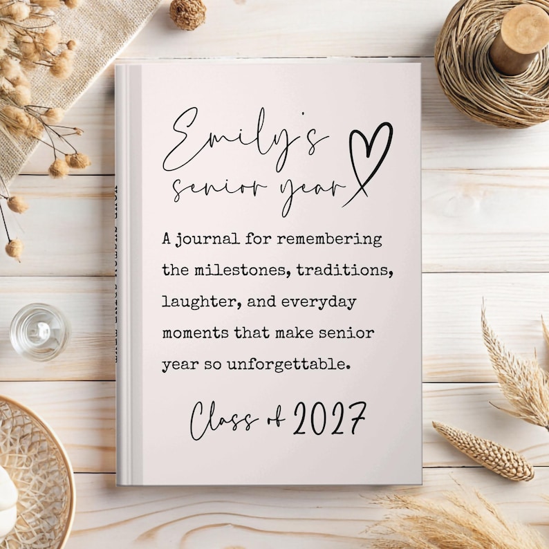 May include: A hardcover journal with the text "Emily's senior year" and "Class of 2027" in black script. The journal's cover includes a heart symbol and a description of the journal's purpose: to remember milestones, traditions, and everyday moments.