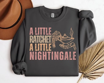 Op de afbeelding: Donkergrijze sweatshirt met de tekst "A LITTLE RATCHET A LITTLE NIGHTINGALE" in roze, beige en perzik. Het ontwerp bevat een skelet hand die een toverlamp vasthoudt. Een bruine hoed staat in de linkerbovenhoek.