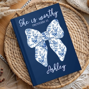 May include: A navy blue journal with a white and blue floral bow design. The text "She is worthy Proverbs 31" is printed on the front cover. The name "Ashley" is printed below the bow.