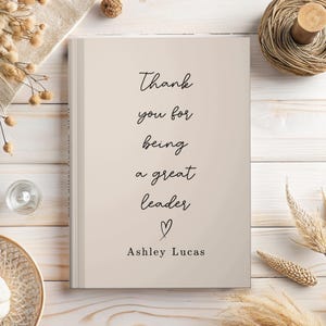 May include: A beige hardcover book with black text that reads Thank you for being a great leader followed by a small heart symbol and the name Ashley Lucas. The book is centered on a light wood surface with dried floral accents nearby.