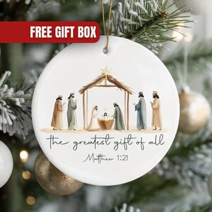 May include: Round white Christmas ornament with a Nativity scene illustration. The image depicts figures around a manger, with the text "the greatest gift of all" and Matthew 1:21. A gold string is attached for hanging.