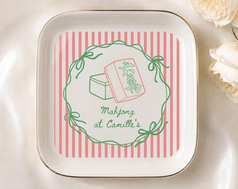 May include: A square white ceramic trinket dish with a gold-toned rim. The center features a pink and white vertical striped background, a green hand-drawn wreath, two mahjong tiles, and the text "Mahjong at Camille's" in green script.
