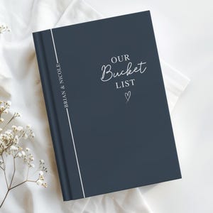 Pu&ograve; includere: Un diario blu navy con le parole "OUR Bucket LIST" in corsivo bianco, un piccolo cuore e i nomi "BRIAN & NICOLE" in verticale lungo il dorso. Il diario &egrave; su una superficie bianca con accenti floreali.