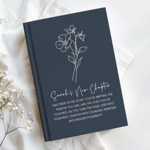 May include: A navy blue journal with the text "Sarah's New Chapter" and a floral line drawing. The journal's cover includes inspirational text about pride, courage, and endless possibilities.