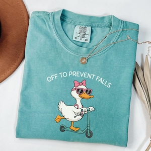May include: Teal Comfort Colors t-shirt with the phrase "OFF TO PREVENT FALLS" and a cartoon duck wearing sunglasses and a pink bow riding a scooter. A gold necklace and a brown hat are also visible.