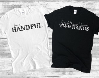 Könnte beinhalten: Zwei T-Shirts mit passendem Design. Das weiße T-Shirt sagt "I'm a handful". Das schwarze T-Shirt sagt "Good thing I have two hands".