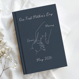 May include: A navy blue journal featuring white text and a line drawing of a parent's hand holding a child's hand. The text reads "Our First Mother's Day," "Mommy," "Emma," and "May 2026."