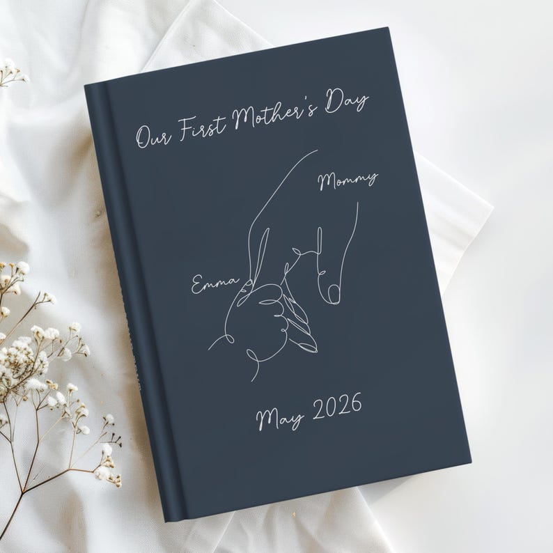 May include: A navy blue journal featuring white text and a line drawing of a parent's hand holding a child's hand. The text reads "Our First Mother's Day," "Mommy," "Emma," and "May 2026."