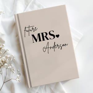 May include: A beige hardcover book featuring black text that reads future MRS. followed by a small heart icon and the name Anderson in a cursive script. The cover has a smooth, matte finish and is positioned at an angle on a white fabric surface.