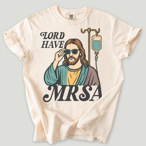 May include: Cream-colored t-shirt featuring a graphic of a person wearing sunglasses, with long hair and a beard, and an IV drip. The text reads "LORD HAVE MRSA" in a vintage font. The shirt has rolled-up sleeves.
