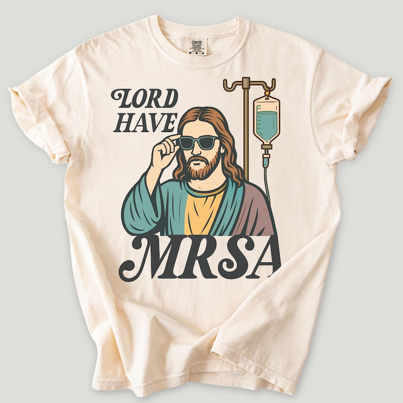May include: Cream-colored t-shirt featuring a graphic of a person wearing sunglasses, with long hair and a beard, and an IV drip. The text reads "LORD HAVE MRSA" in a vintage font. The shirt has rolled-up sleeves.