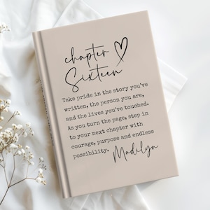 May include: A beige hardcover journal with the words "chapter Sixteen" and a heart symbol in black script. The journal includes inspirational text and the name "Madilyn" in cursive. The journal is on a white surface.