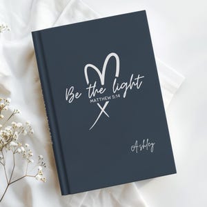 May include: A navy blue journal with the text "Be the light" and "Matthew 5:14" in white script. A white heart-shaped design and the name "Ashley" are also featured. The journal is lying on a white surface.