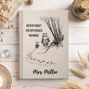 May include: A light beige hardcover book featuring a black line drawing of Winnie the Pooh and Piglet walking away. The text reads, "NEVER FORGET THE DIFFERENCE YOU MAKE." and "Mrs. Miller" is printed in a black script font at the bottom.