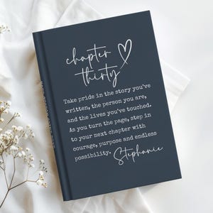 May include: A navy blue journal with the words "chapter thirty" and a heart symbol in white script. The journal includes inspirational text and the name "Stephanie" in cursive. The journal is on a white cloth.