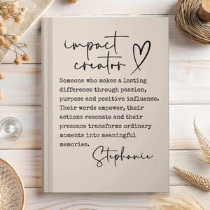 May include: A beige-colored book with the words "impact creator" and a heart symbol in black script. The text below describes someone who makes a lasting difference. The name "Stephanie" is written at the bottom.
