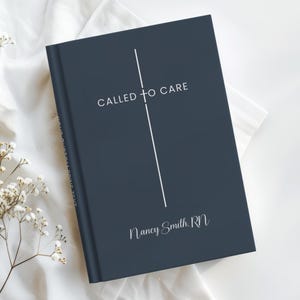 May include: A navy blue journal with the text "CALLED TO CARE" and "Nancy Smith, RN" in white script. A vertical white line intersects the text. The journal is on a white surface with delicate white flowers.