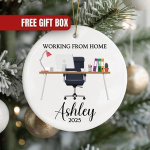 May include: A white ceramic Christmas ornament with a home office design. The ornament features a desk with a computer, chair, and desk accessories. Text on the ornament reads "WORKING FROM HOME" and "Ashley 2025". A red banner at the top reads "FREE GIFT BOX".