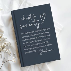 May include: A navy blue journal with the words "chapter seventy" and a heart symbol in white script. The journal includes inspirational text and a signature. The journal is on a white surface.