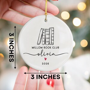 May include: A round white ceramic ornament featuring a black line drawing of four books, the text WILLOW BOOK CLUB, the name olivia, a small red heart, and the year 2026. The ornament measures 3 inches by 3 inches and includes a gold hanging cord.