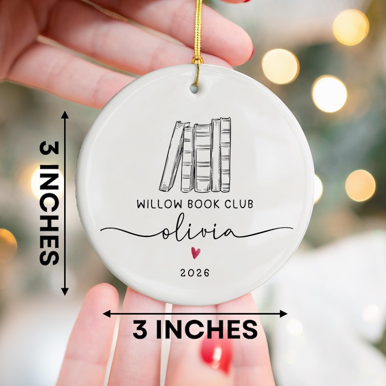 May include: A round white ceramic ornament featuring a black line drawing of four books, the text WILLOW BOOK CLUB, the name olivia, a small red heart, and the year 2026. The ornament measures 3 inches by 3 inches and includes a gold hanging cord.