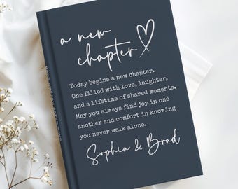 May include: A dark blue journal with white script reading "a new chapter" and a heart symbol. The journal contains a message about love and shared moments, signed "Sophia & Brad".