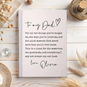 May include: A white journal with the text "To my Dad" and a heart symbol. The journal contains a heartfelt message from "Olivia" expressing gratitude and love. The journal is on a light wooden surface with decorative elements.