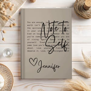 May include: A beige journal with a motivational quote about self-love and a heart with the name Jennifer written in cursive.
