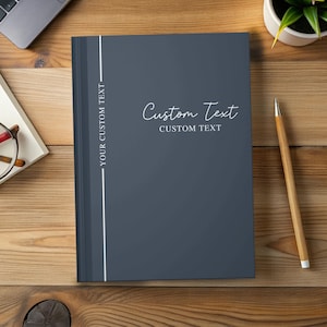 May include: A dark blue notebook with the words "Custom Text" in a cursive font. The notebook has a white vertical stripe on the left side with the text "YOUR CUSTOM TEXT". A pencil rests on a wooden surface.