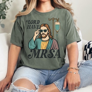 May include: Olive green t-shirt featuring a graphic of Jesus wearing sunglasses, with the words "LORD HAVE MRSA". The image includes an IV drip bag. The shirt is a casual, unisex style.