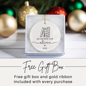 May include: A round white ceramic ornament featuring a black line drawing of four books, the text "WILLOW BOOK CLUB", the name "olivia" in script, a small red heart, and the year "2026". The ornament is displayed inside a white gift box.