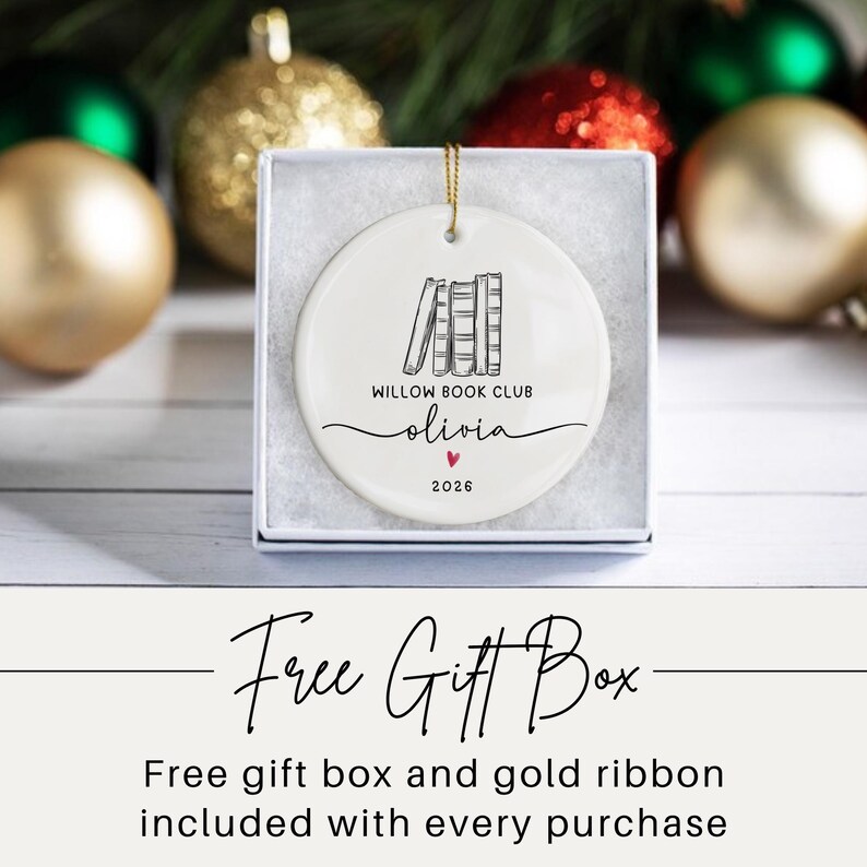May include: A round white ceramic ornament featuring a black line drawing of four books, the text "WILLOW BOOK CLUB", the name "olivia" in script, a small red heart, and the year "2026". The ornament is displayed inside a white gift box.