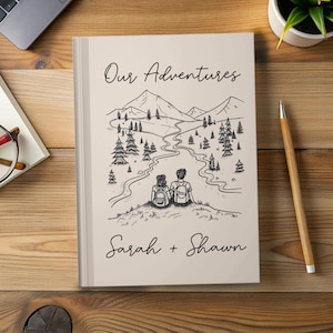 May include: A beige journal with the words "Our Adventures" and the names "Sarah + Shawn" in script. The cover features a black and white illustration of a couple with backpacks overlooking a mountain landscape with a river and trees.