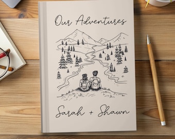 May include: A beige journal with the words "Our Adventures" and the names "Sarah + Shawn" in script. The cover features a black and white illustration of a couple with backpacks overlooking a mountain landscape with a river and trees.
